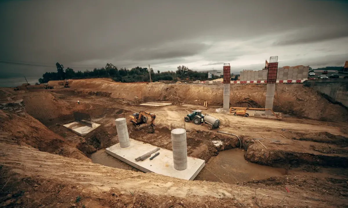 A wide excavation site with poured concrete columns, an excavator and a tractor under a gray sky.