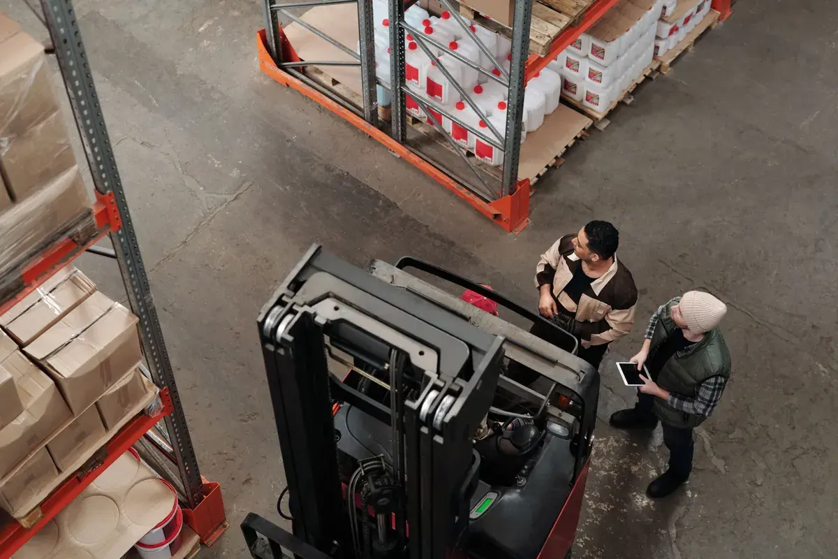 Overhead view of two warehouse workers with a tablet standing beside a forklift among stocked pallet racks.