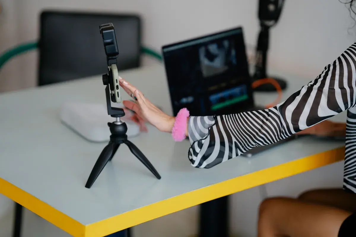 A hand adjusts a phone on a small tripod beside a laptop showing a video editing timeline.