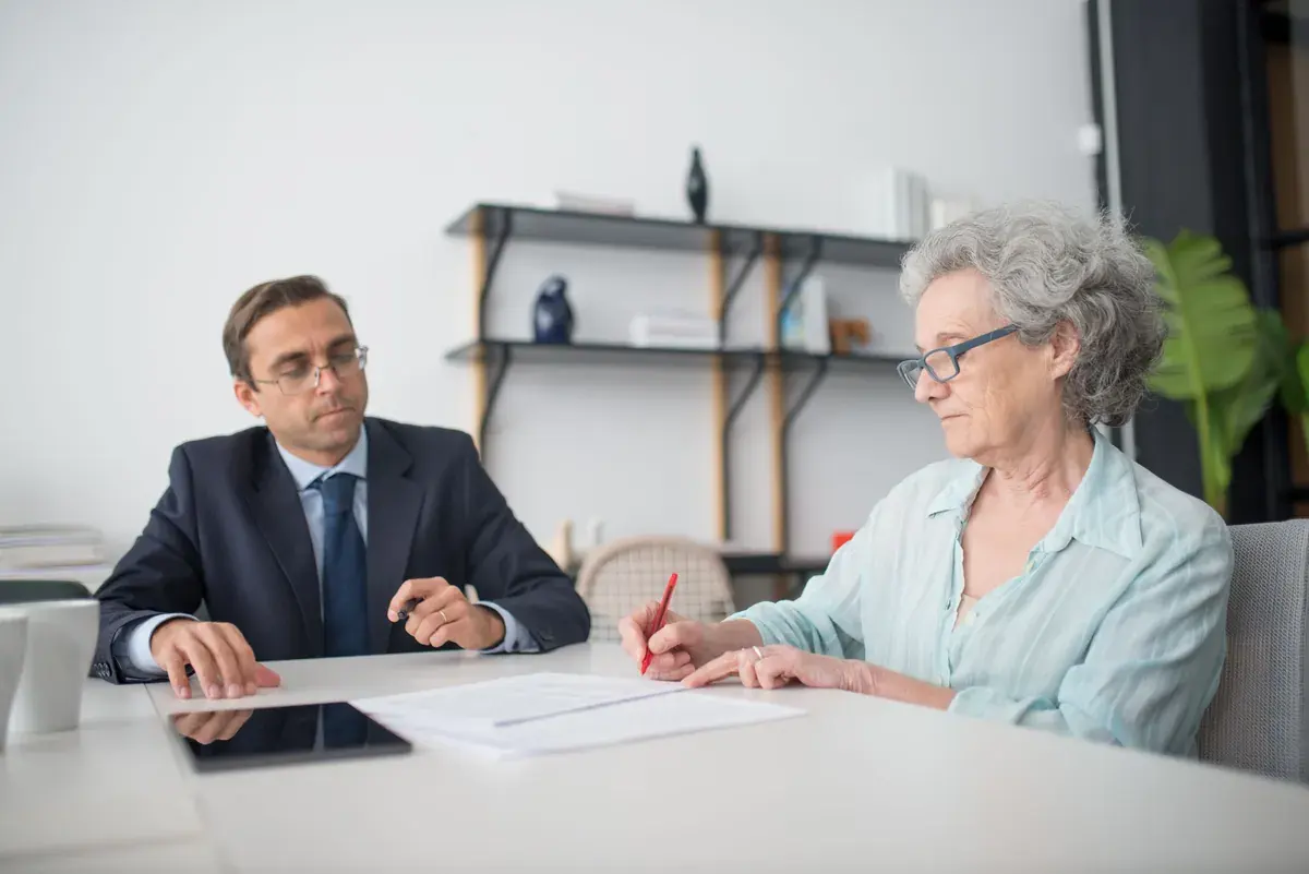 An older woman signs a document while a man in a suit sits beside her.