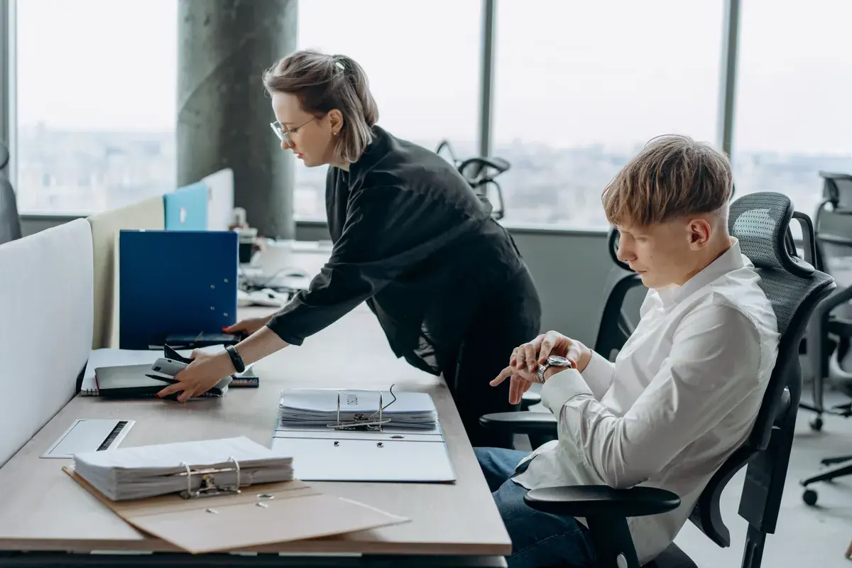 Two colleagues review open ring binders and a monitor at an office workstation