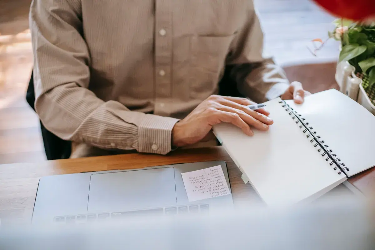 A person in a linen shirt turns the page of a spiral notebook beside an open laptop.