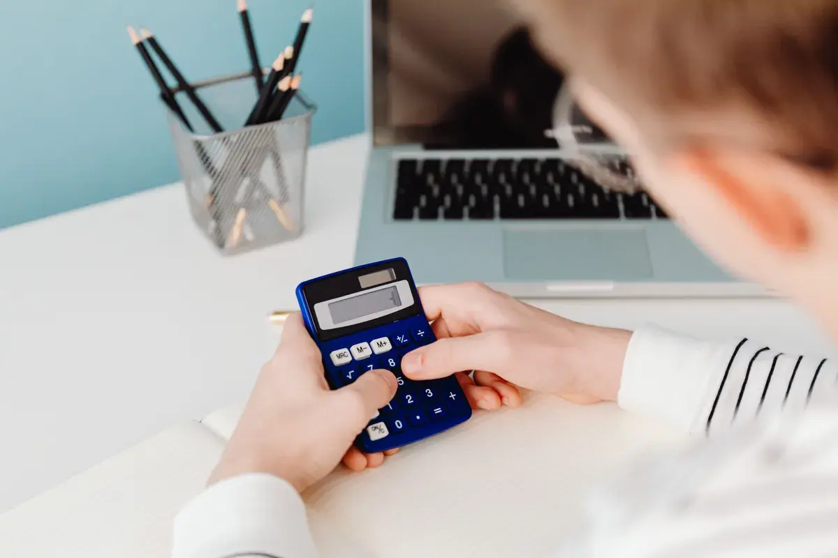 Close-up of hands pressing keys on a blue calculator over a notebook next to a laptop.