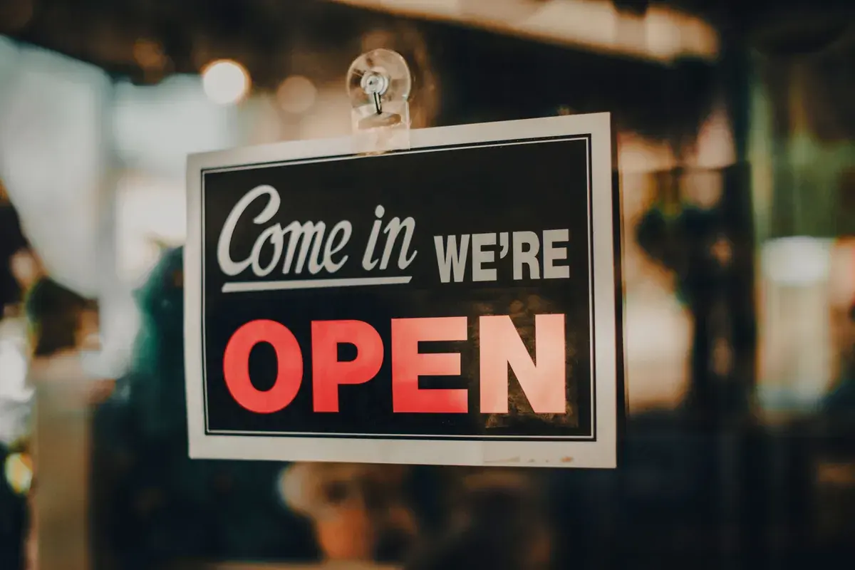 A come in we are open sign hangs in the glass door of a small shop.