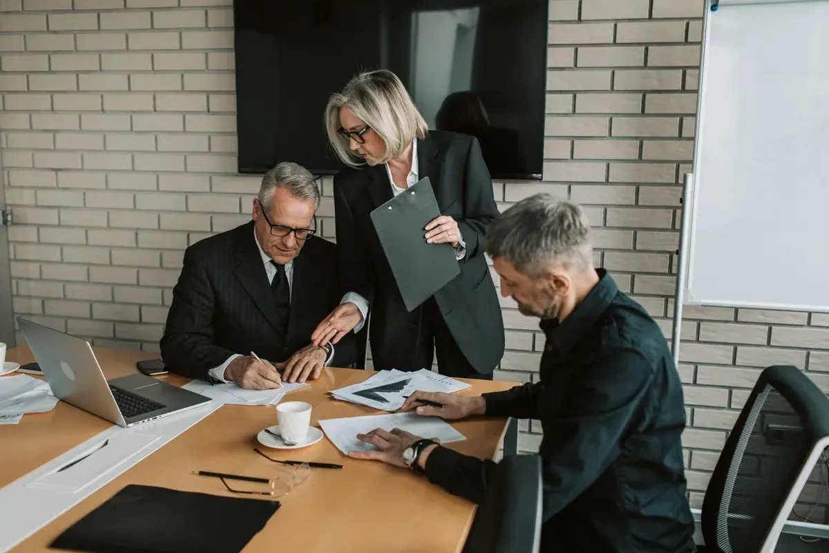 Three professionals in an office review and sign papers spread across a wide table.
