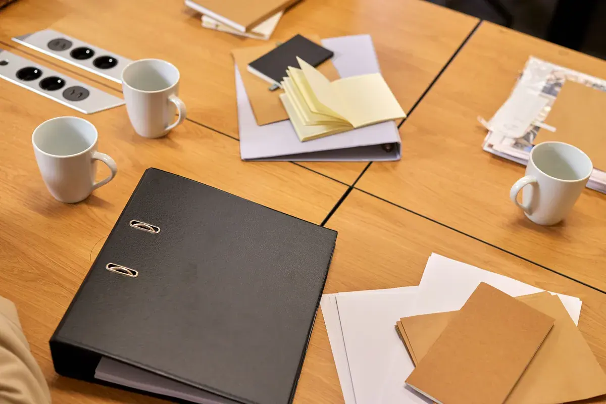 Ring binders, notebooks and loose papers spread across a meeting table with coffee mugs.
