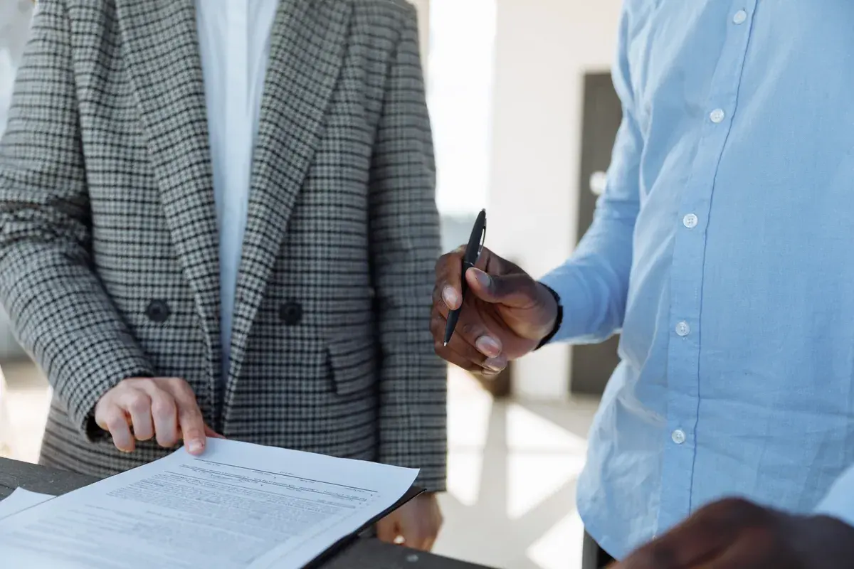 Two professionals stand at a desk reviewing a printed multipage document while one holds a pen.