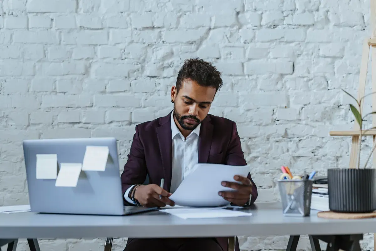 A business owner at a desk reading printed pages beside a laptop in a bright office