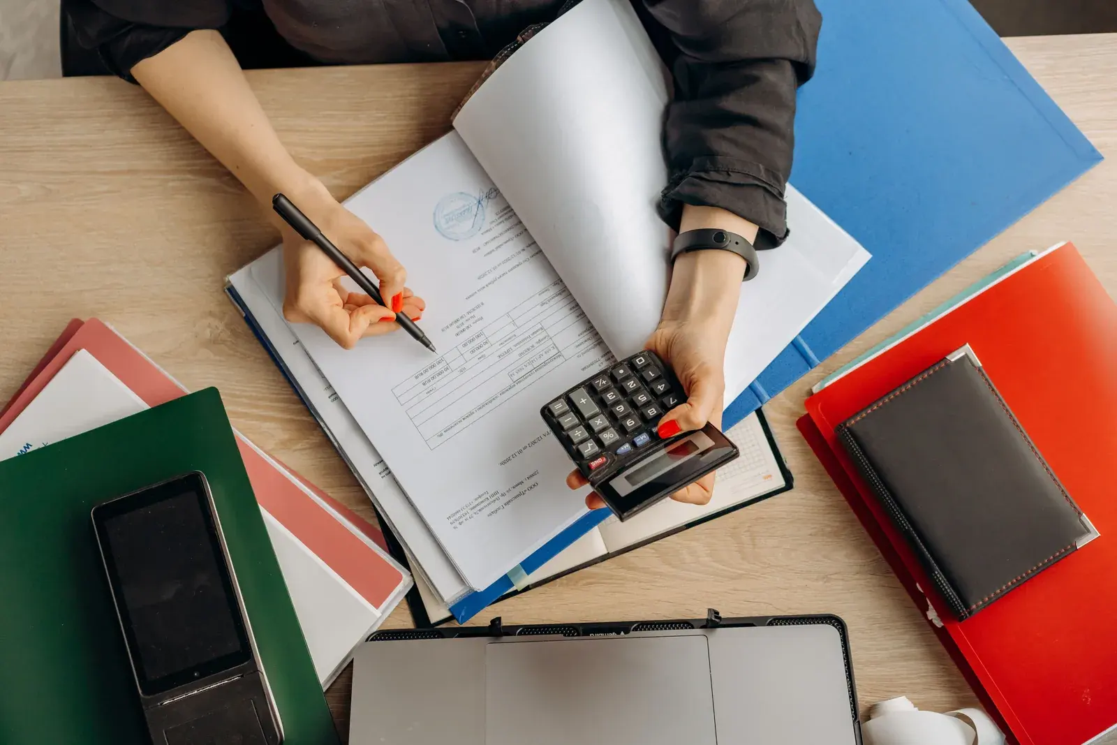 Overhead view of an owner holding a calculator over an open ledger surrounded by colored binders