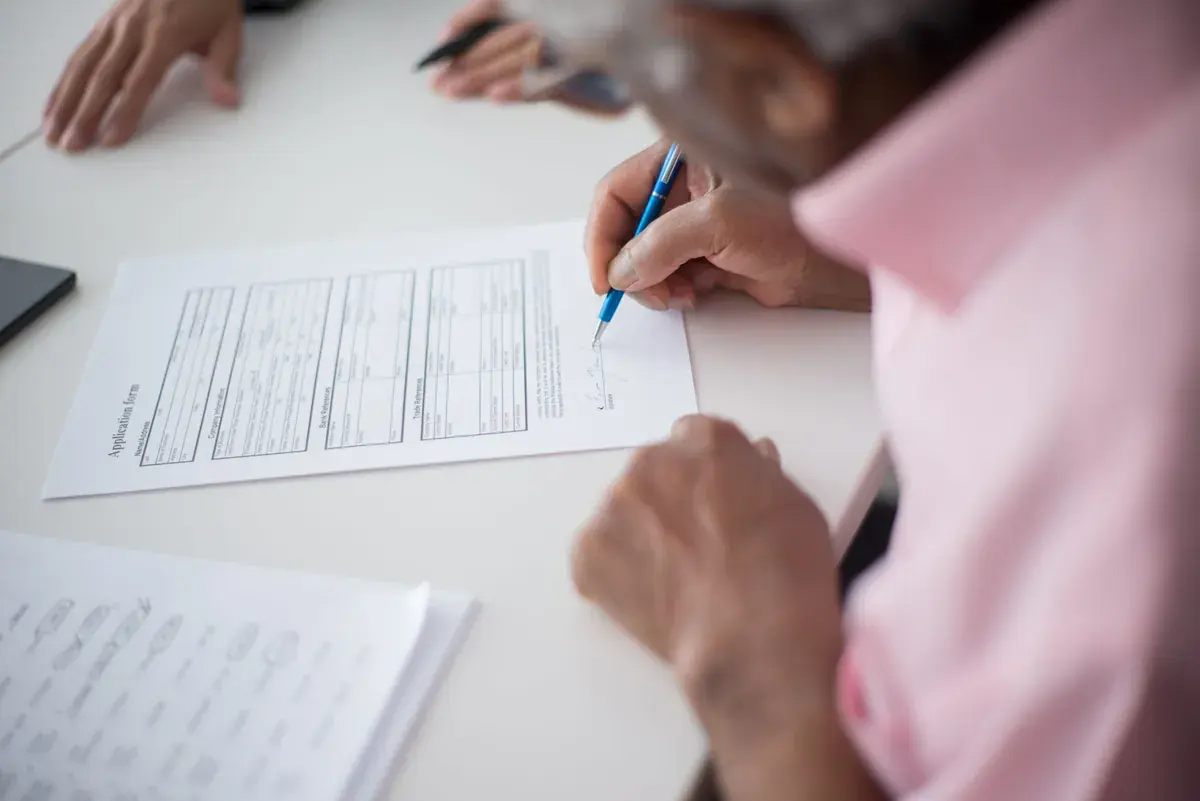 An older adult signs a printed form while another person helps across the table