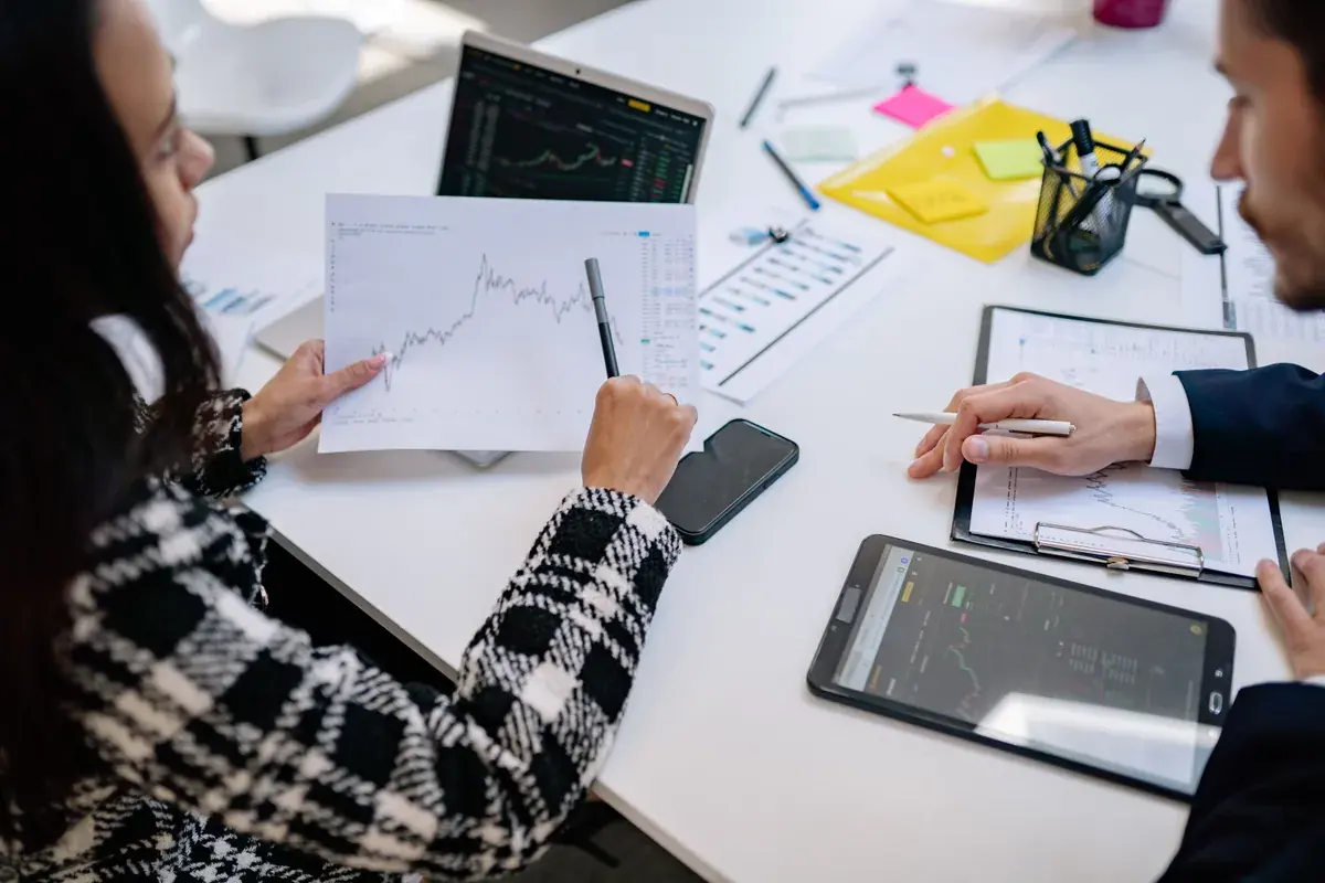 Two colleagues compare printed performance charts across a bright meeting table with a laptop and tablet
