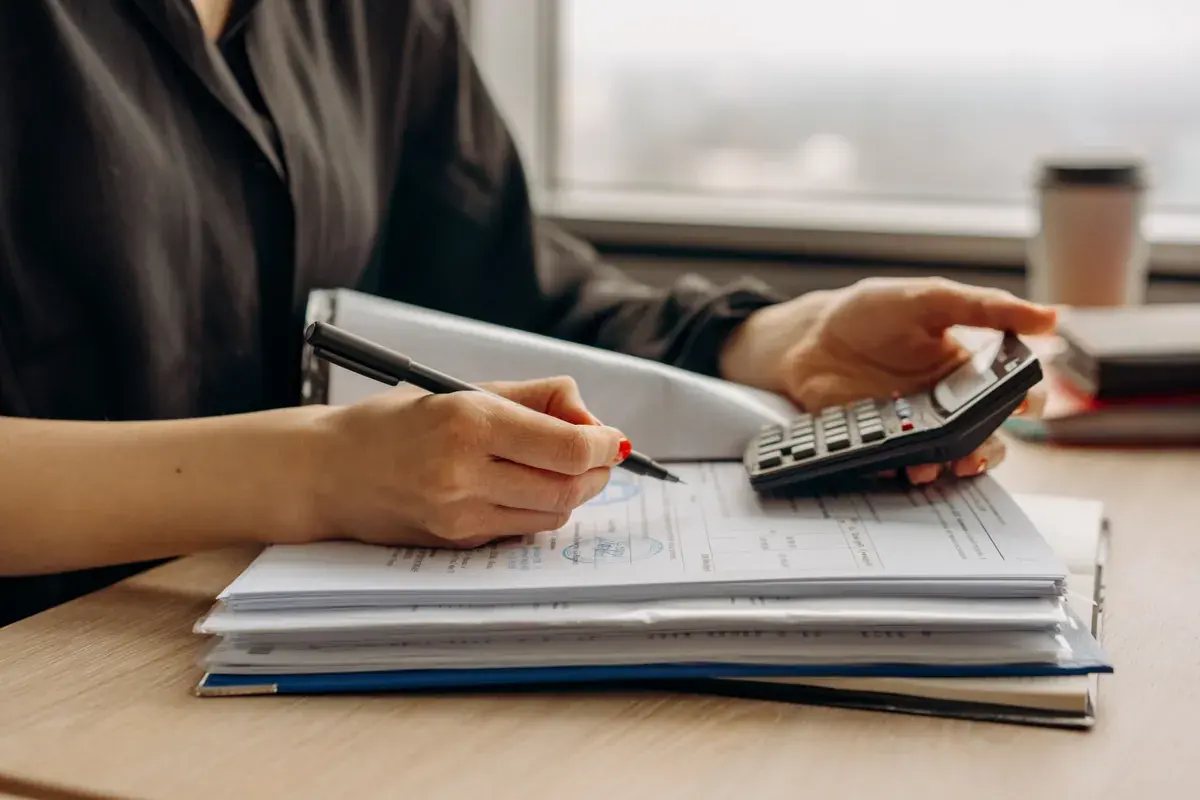 A person at a desk marks up a stack of documents in a binder while working a calculator by a window