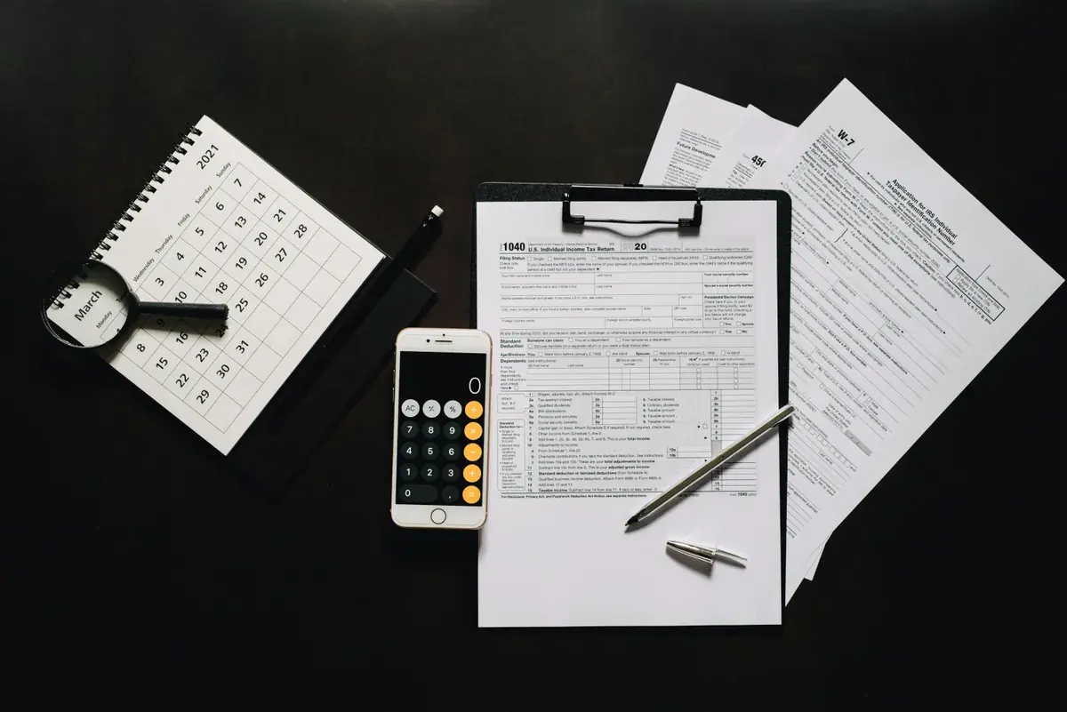 A tax return form on a clipboard beside a desk calendar, a phone calculator, and pens on a dark desk