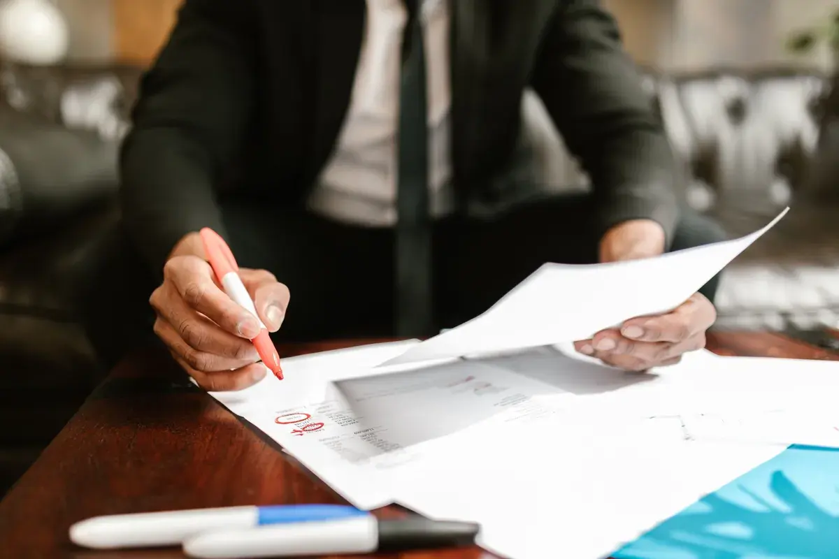 A person in a suit uses a red pen to review printed financial documents on a polished wooden desk