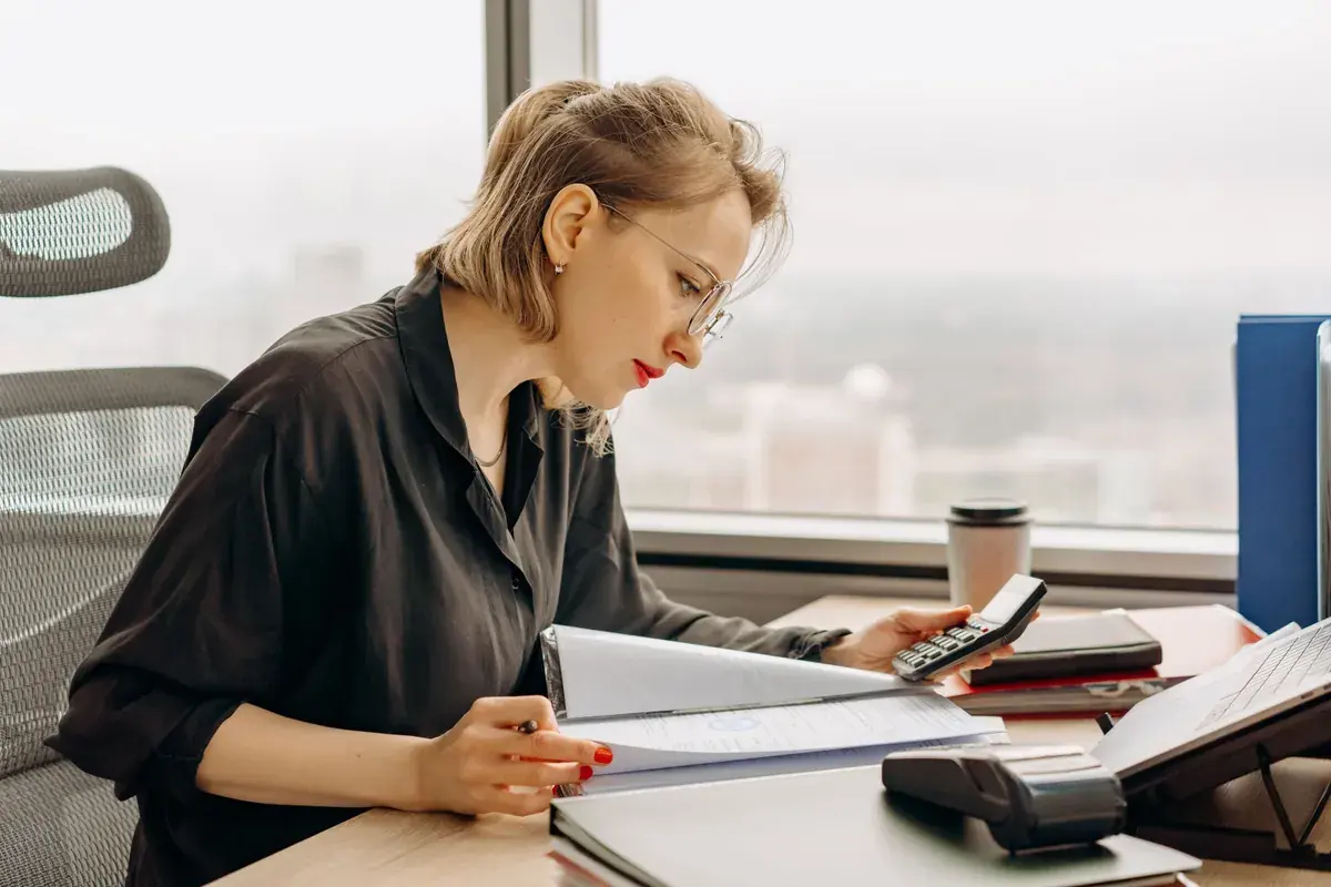 A professional at an office desk uses a calculator while reviewing a stack of printed financial documents by a window