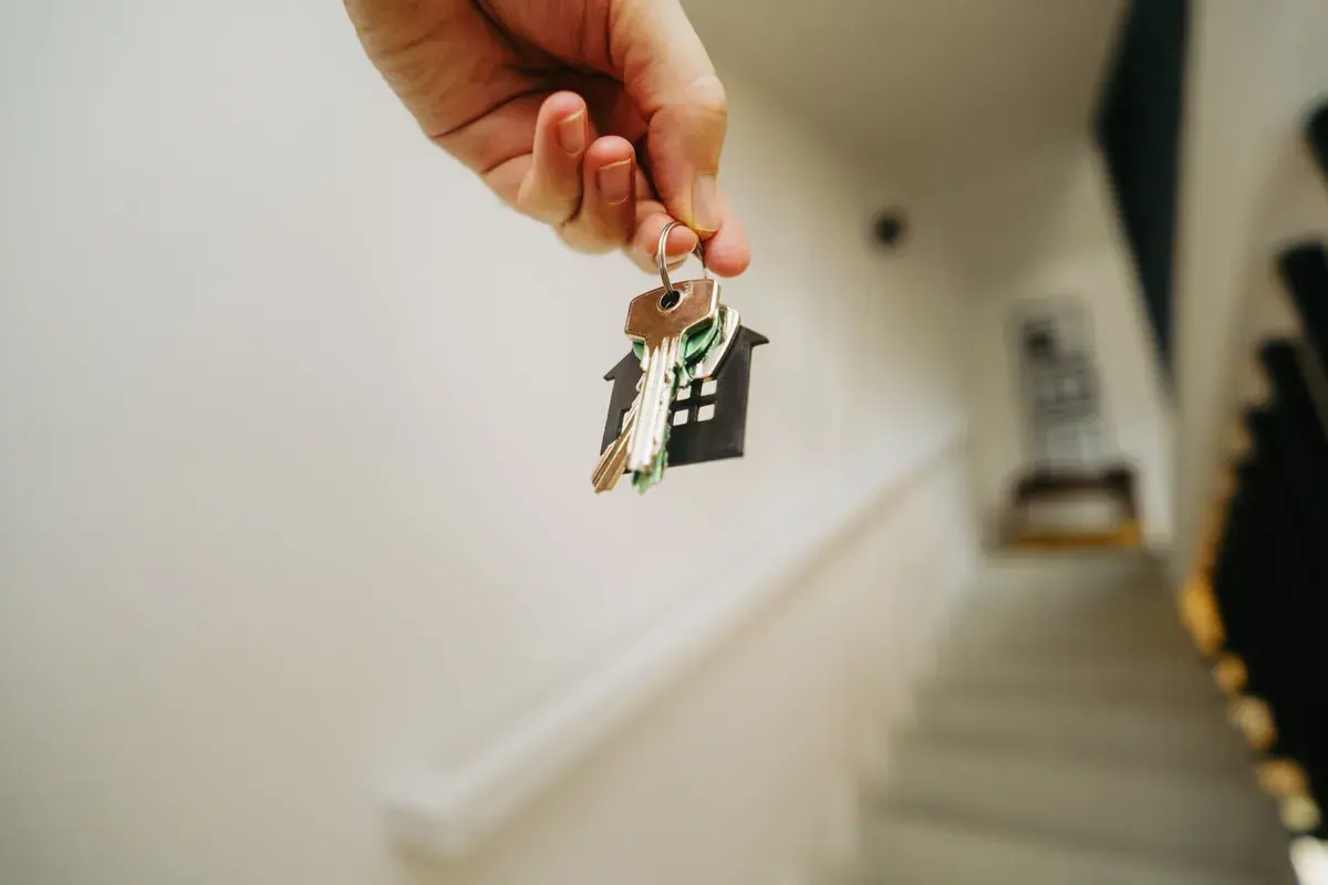 Hand holding a set of keys with a house shaped keychain in a bright hallway