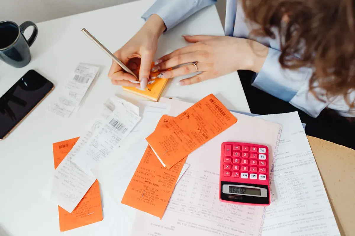 Hands sorting paper receipts beside a calculator and coloured notes on a desk