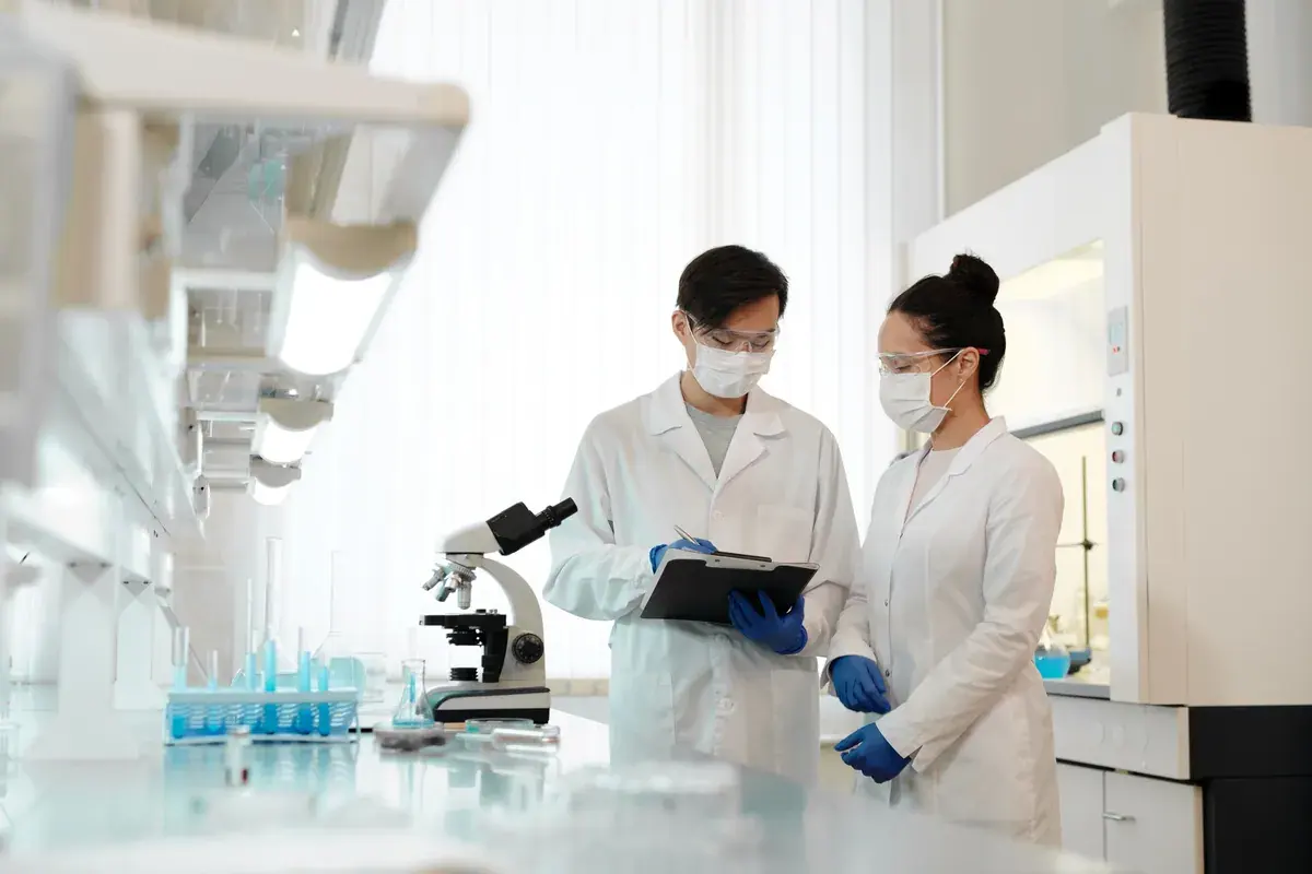 Two scientists in lab coats reviewing notes on a clipboard beside a microscope in a bright lab