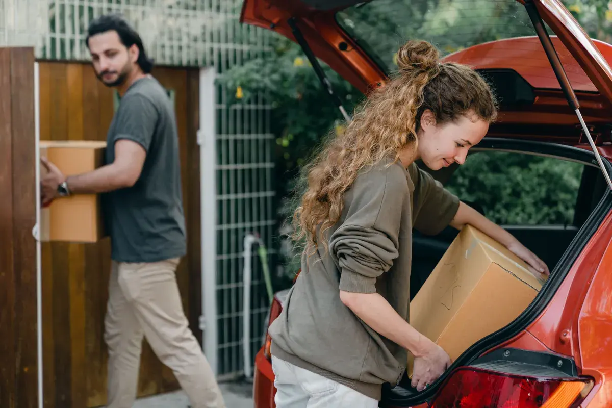 A couple carries cardboard moving boxes from an open car hatch toward a wooden gate on a bright day