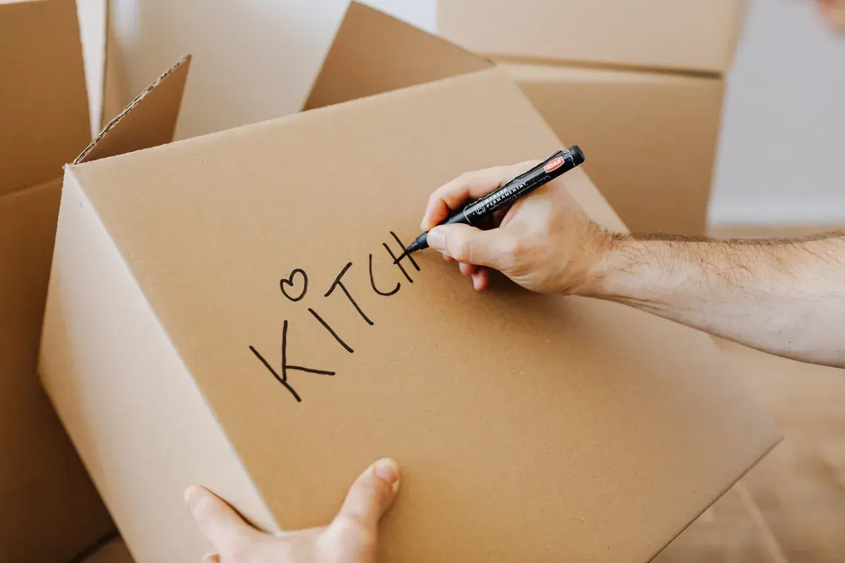 A hand writes the word kitchen with a black marker on top of a sealed cardboard moving box