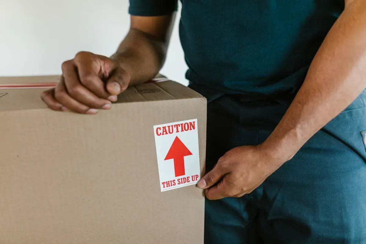 A person steadies a sealed cardboard box with a red arrow label on the side indoors