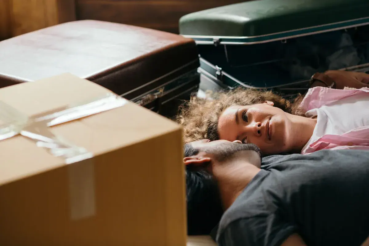 Two people rest together on the floor among stacked vintage suitcases and a cardboard moving box