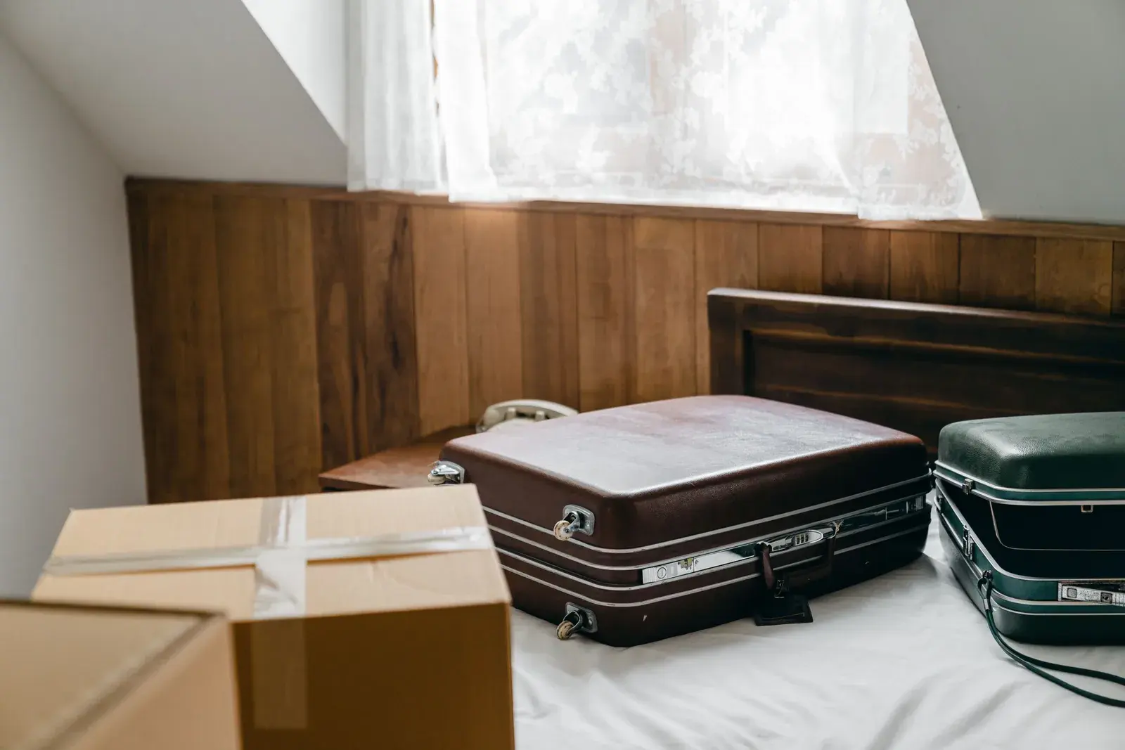 Vintage brown and green leather suitcases on a bed next to a cardboard moving box in a warm wood paneled room