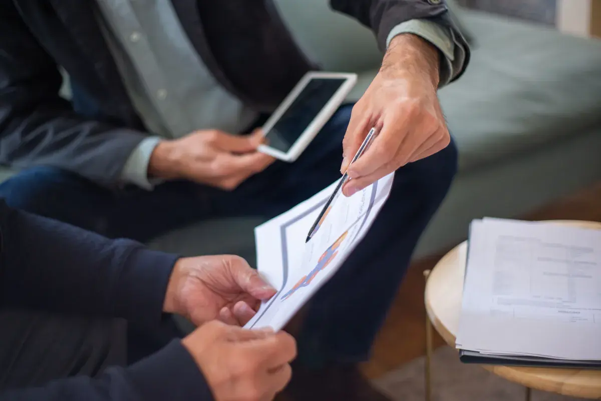 Two people on a couch review a printed report; one hand holds a pen pointing at a chart, the other holds a small phone