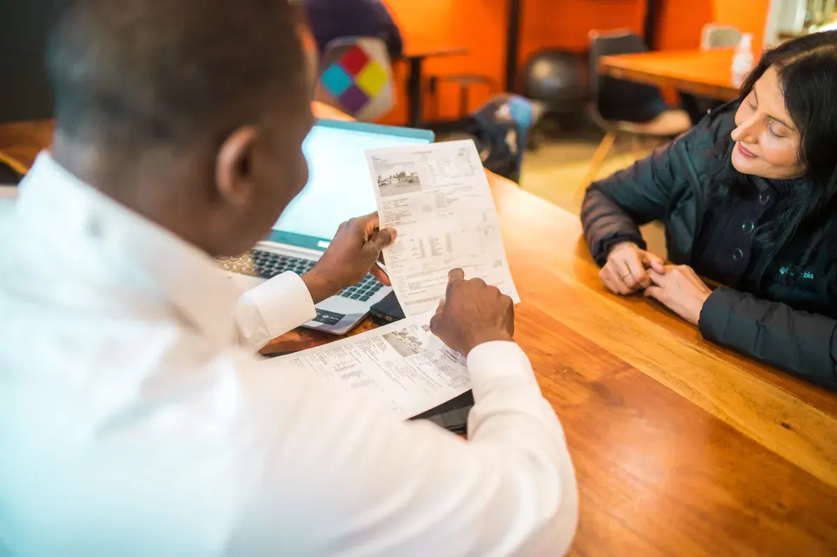 Two adults at a wooden table review a printed multi-page document together beside an open laptop in a warm meeting room
