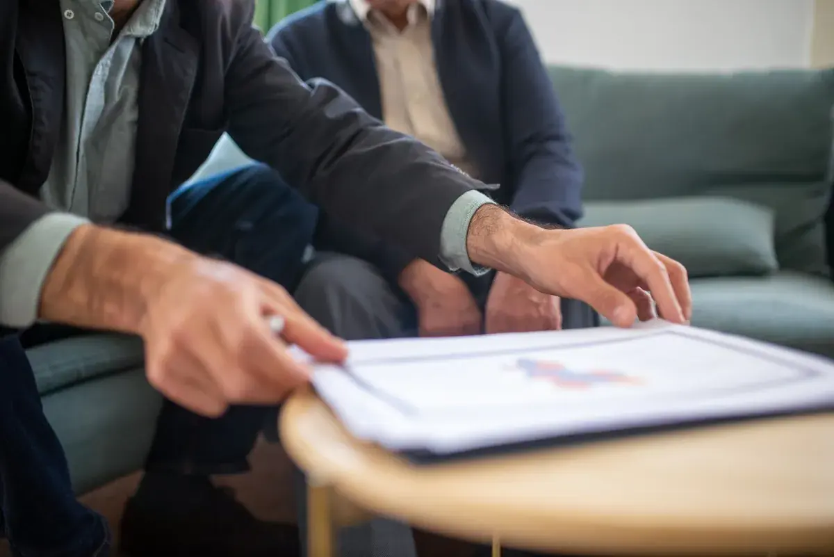 Two people on a green sofa lean over a printed report on a small wooden table, one hand steadying the page, the other pointing