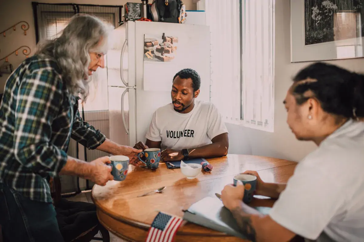 Three diverse volunteers meeting around a small kitchen table with coffee mugs and paperwork