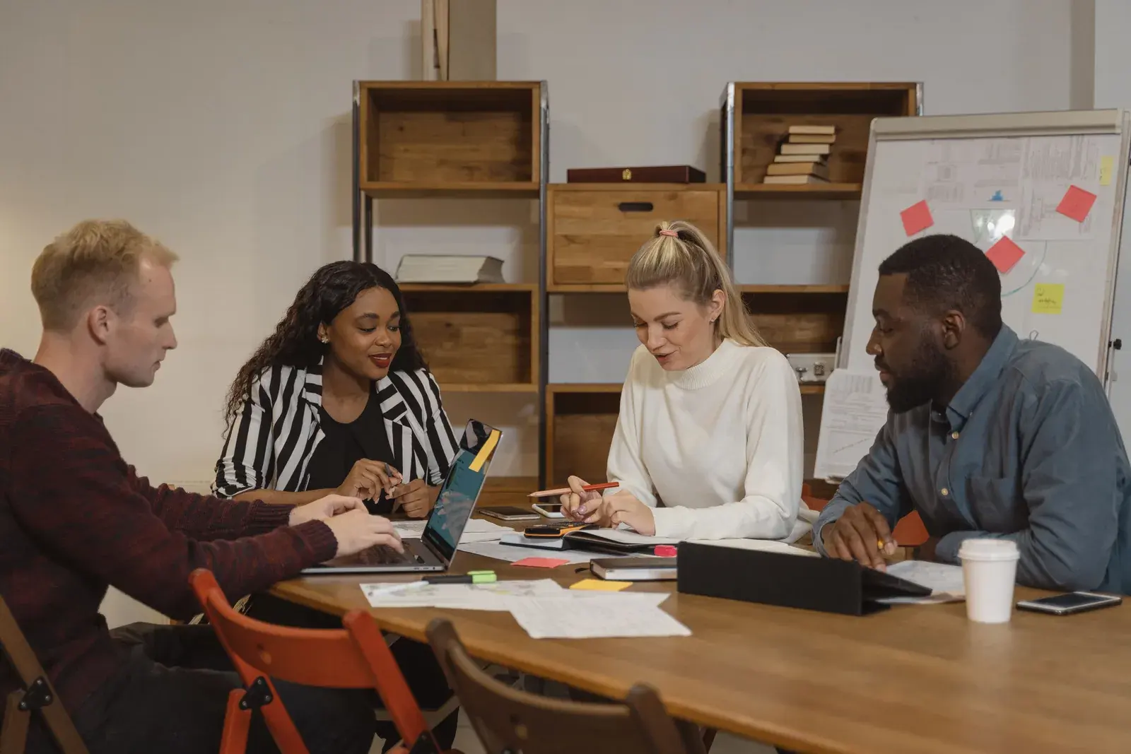 A mixed team of four adults at a wood table collaborating with a laptop, notebooks, and coffee