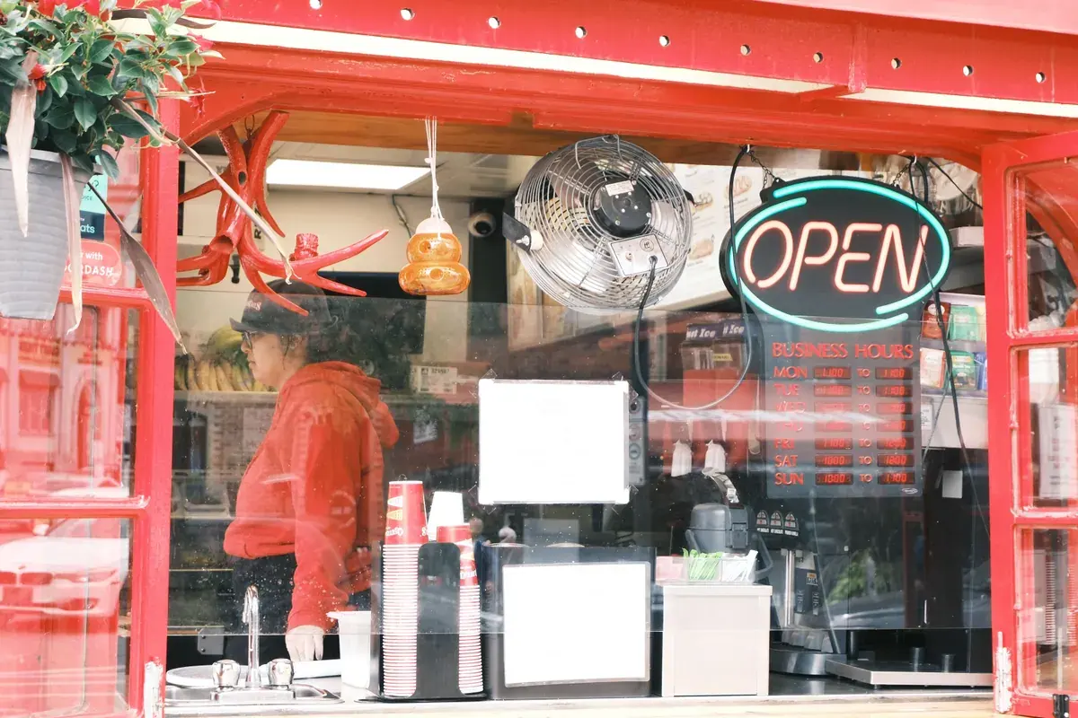 Un pequeño local de comida visto desde la acera, con el letrero de abierto encendido y una persona trabajando detrás del mostrador.