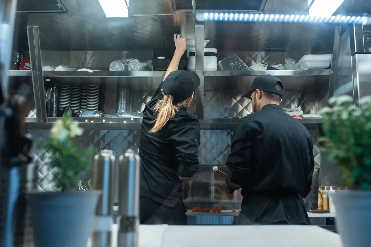 Dos cocineros con uniforme oscuro trabajan uno al lado del otro en la línea de una cocina comercial de acero inoxidable.