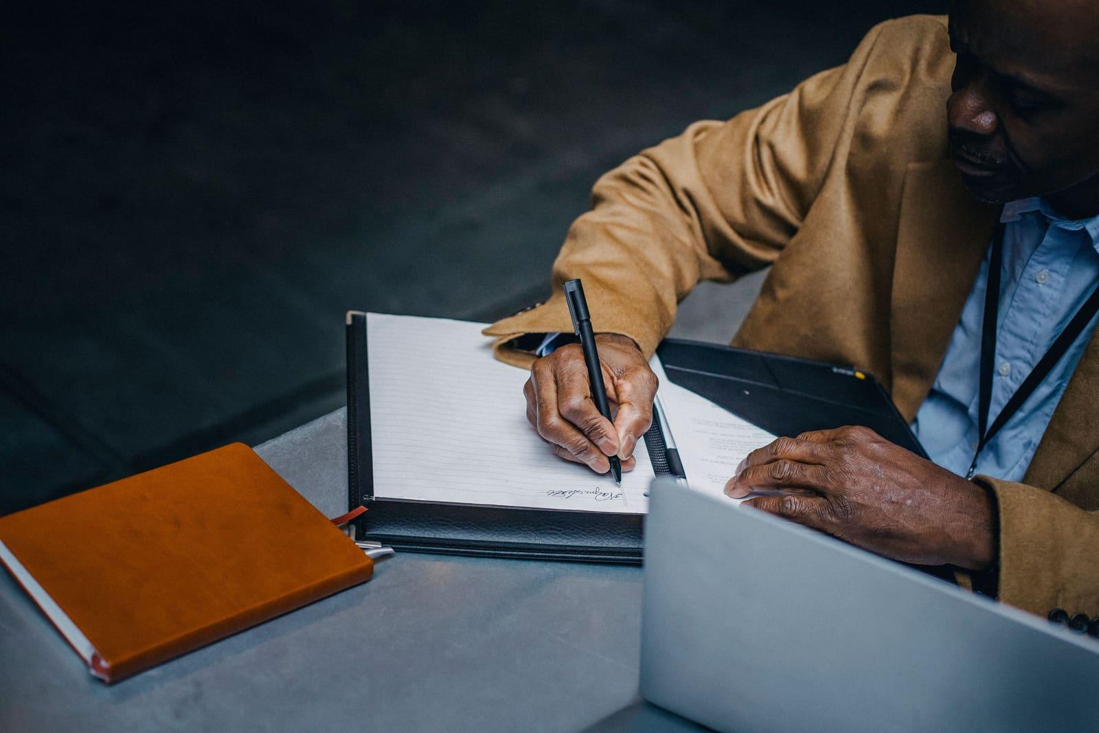 A professional writing notes in a planner beside a laptop at a desk