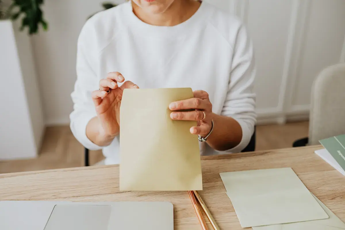 A person seals a kraft paper envelope at a light wooden desk.