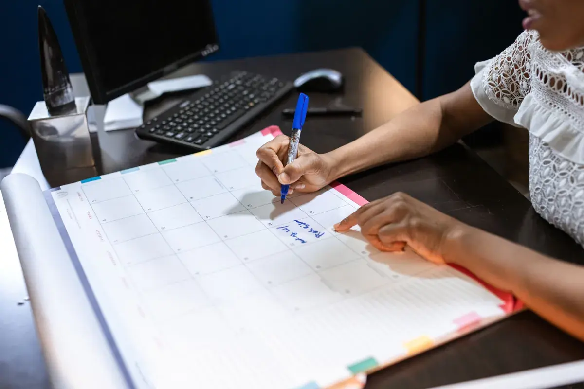 A hand writes a reminder on a large paper desk calendar next to a keyboard.