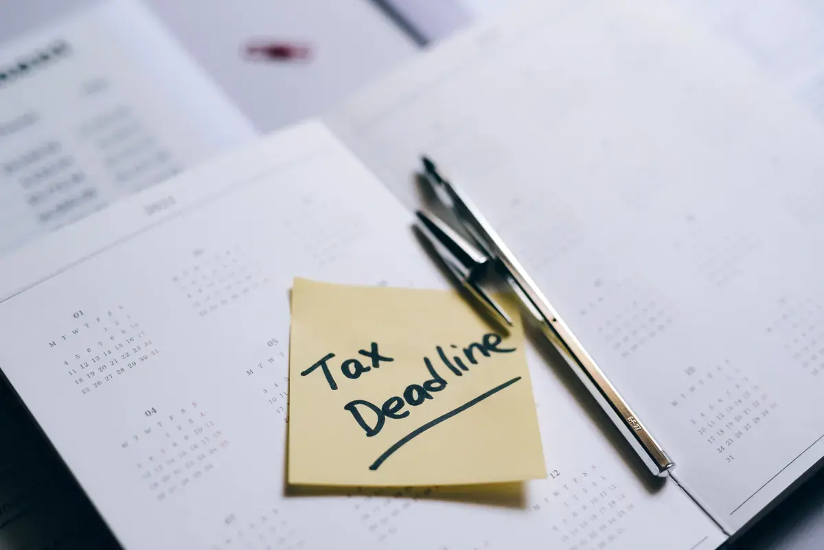 A yellow sticky note with a tax deadline reminder rests on an open paper planner beside a slim pen.