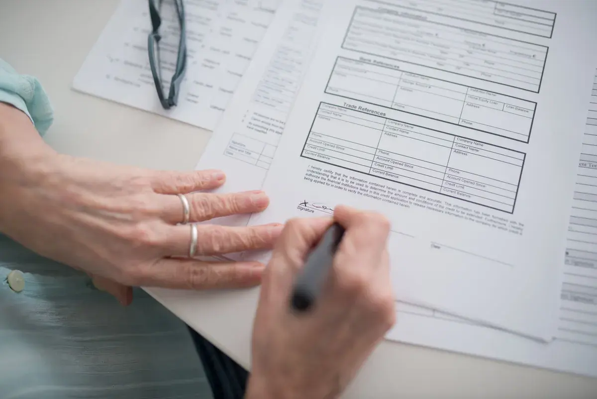 Hands sign a multi-page business form at a desk beside reading glasses and other paperwork.