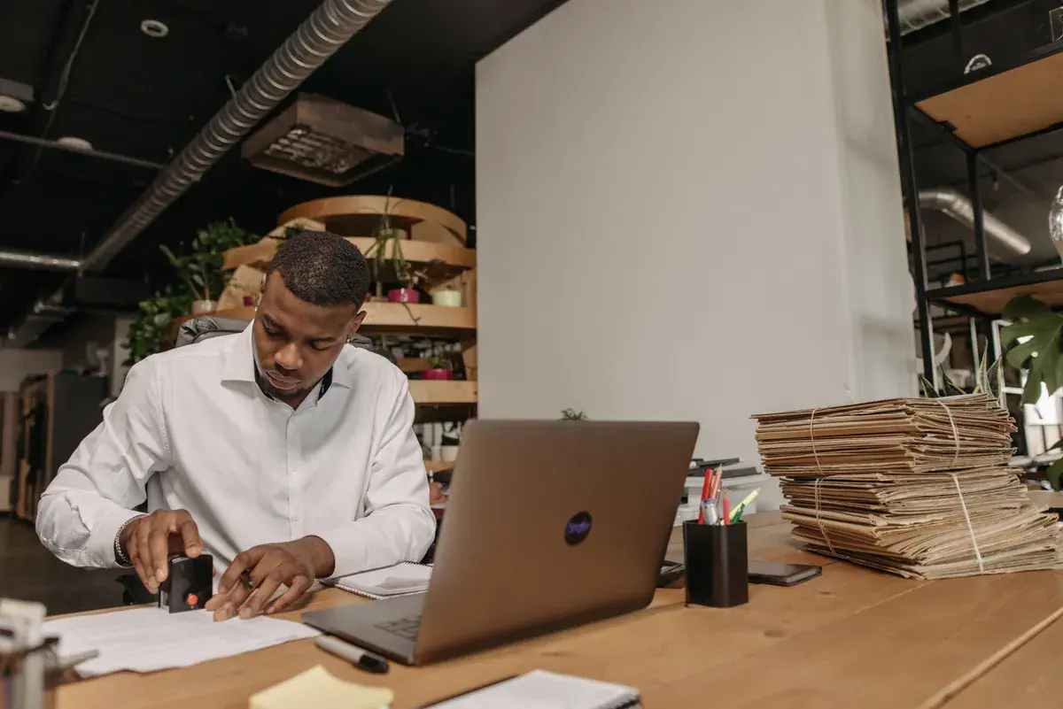 A man stamps a document at a desk with a laptop and stacks of tied folders.