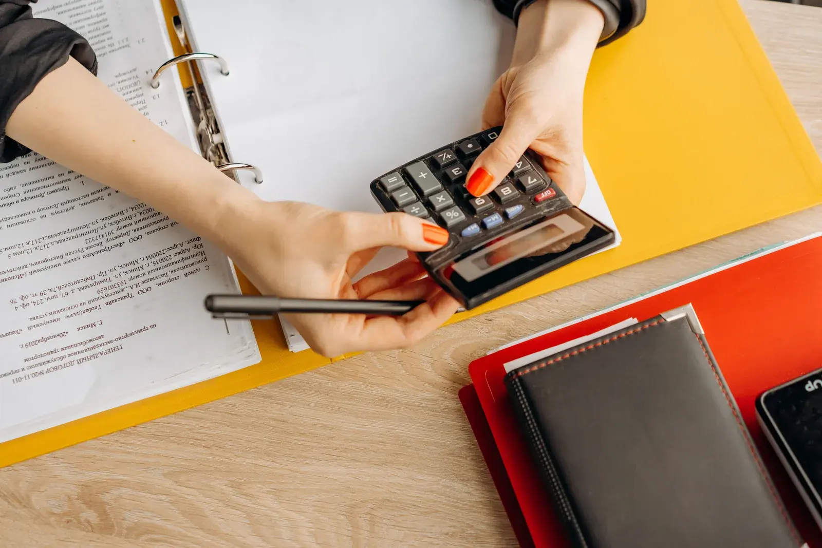 Hands using a calculator at a desk with a stack of documents and a folder within reach