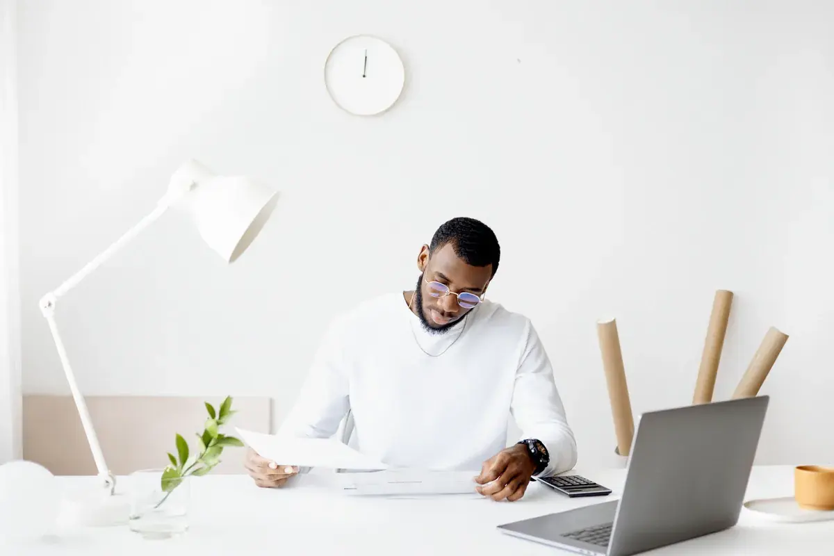 A man at a bright white desk reviews a printed page beside a laptop and calculator.