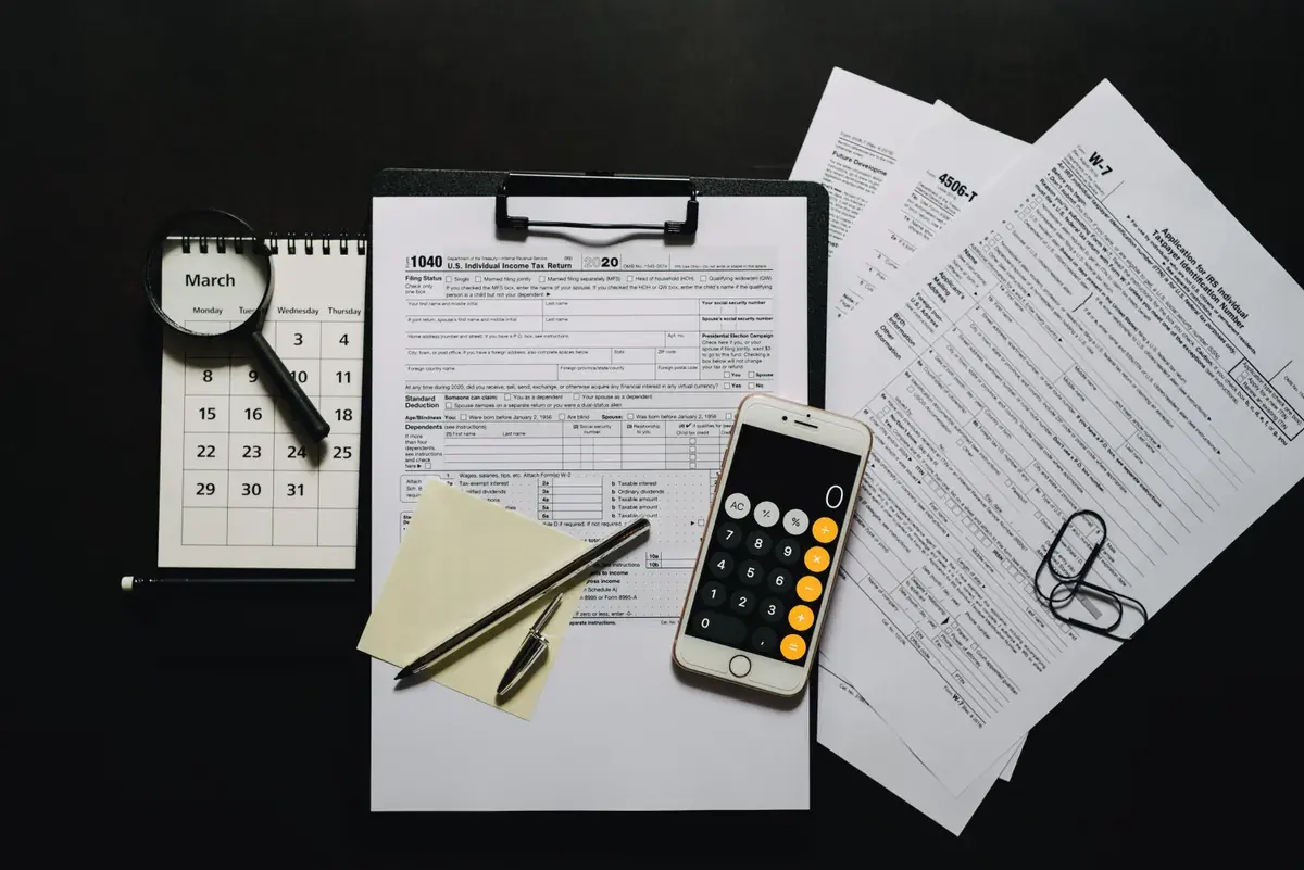 Blank federal tax forms on a clipboard beside a phone calculator, a magnifying glass, and a pen on a dark desk.