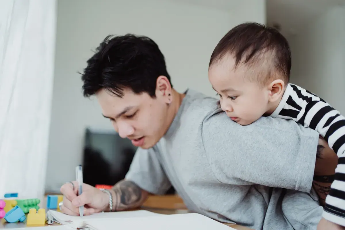 A father writes in a notebook at home while carrying his baby on his shoulder
