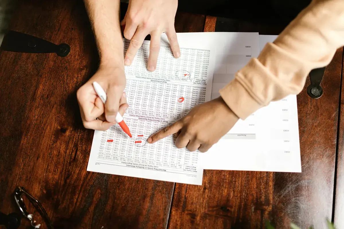 Two people point at rows on a printed table of figures and mark them with a red pen at a wooden desk
