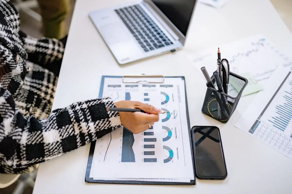 A person marks up a printed chart on a clipboard beside a laptop, phone and pens on a white desk.