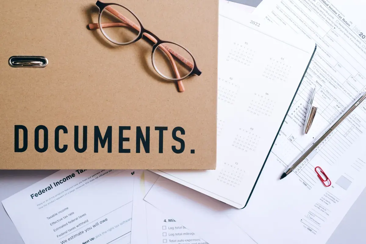 A folder marked documents rests with reading glasses, a calendar page and printed tax return forms on a desk.
