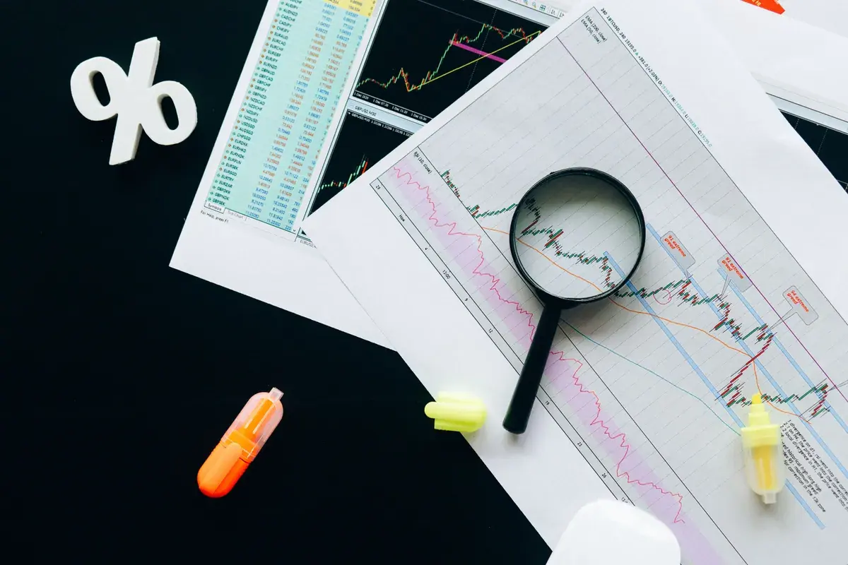 Printed market charts on a dark desk, inspected with a magnifier and marked with highlighters.
