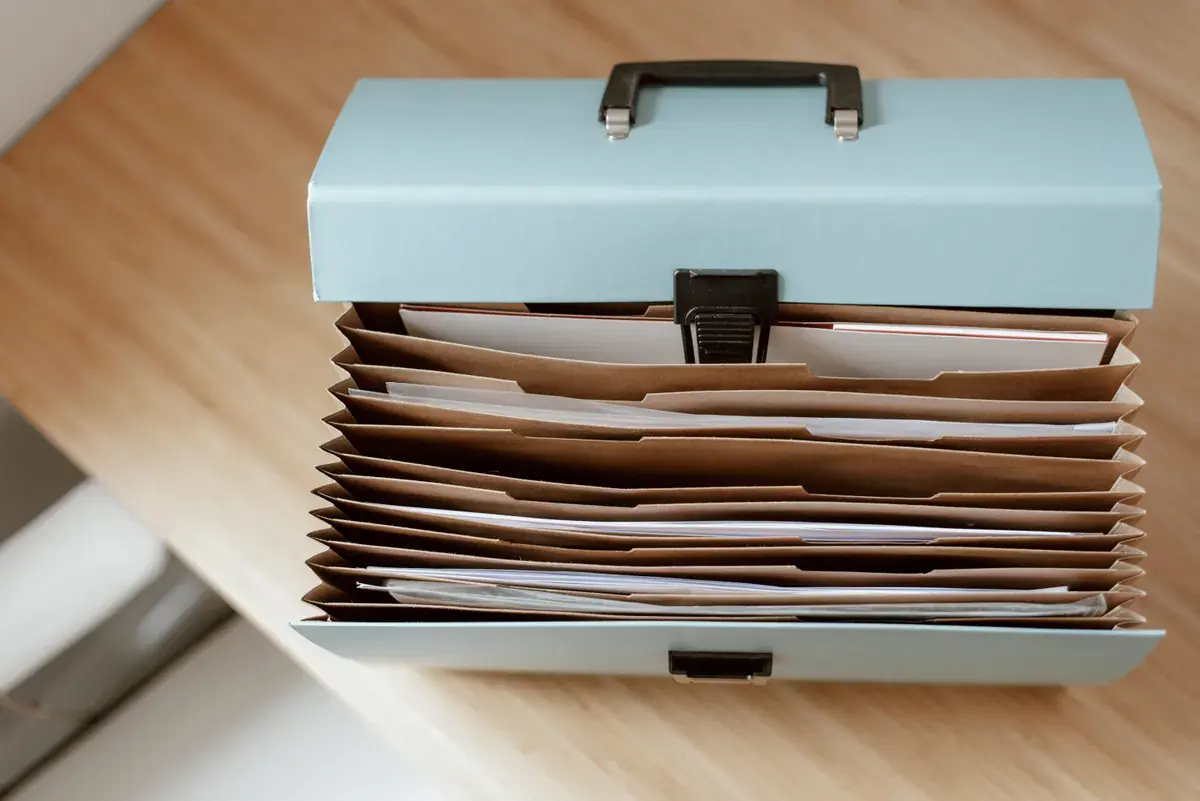 An open accordion file box on a wooden desk holding tabbed dividers and sorted papers