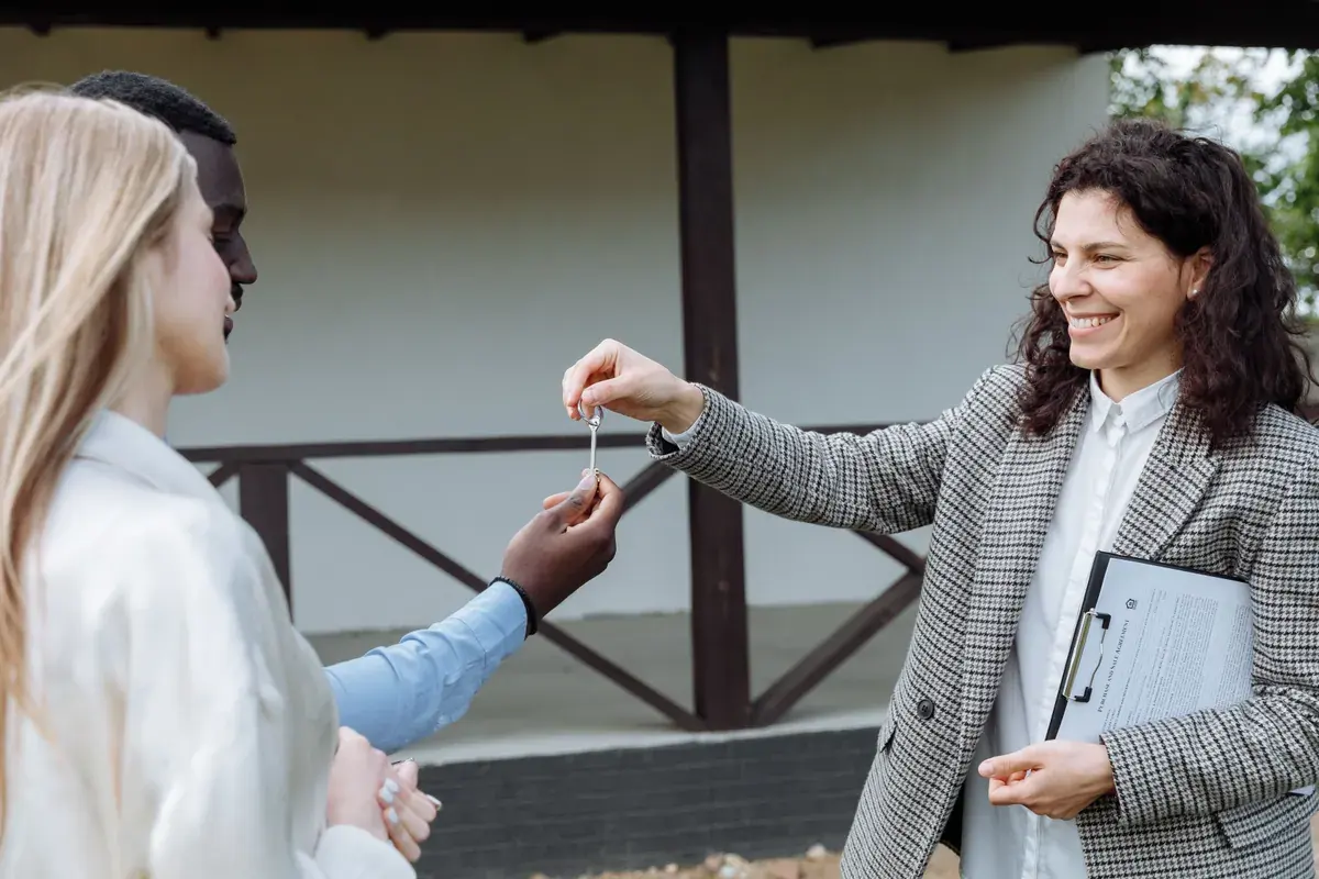A real estate agent hands a set of keys to two people standing outside a home
